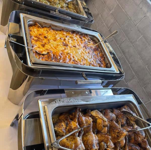 A buffet setup featuring trays of baked lasagna, roasted chicken, and stuffed meatballs in stainless steel serving trays.