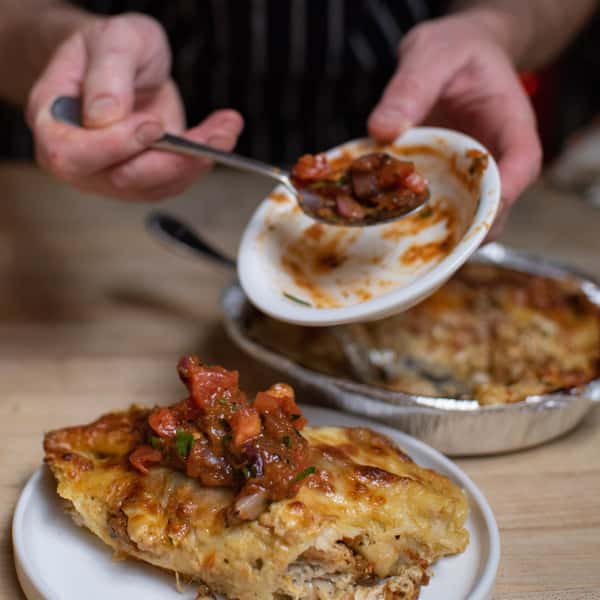 Chef garnishing a slice of cheesy lasagna with fresh tomato salsa, served alongside a foil container of more lasagna, in a rustic kitchen setting.