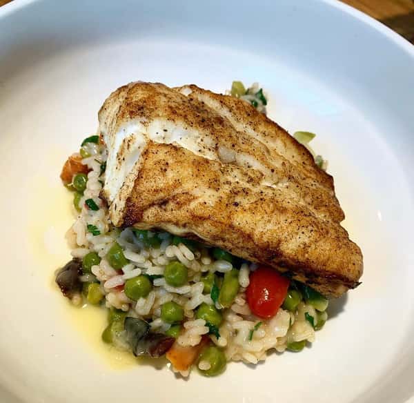 A portion of grilled white fish served over a bed of rice mixed with peas, tomatoes, and herbs, drizzled with a light oil.