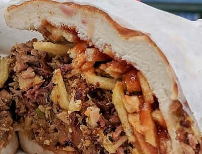 An American and international restaurant-style sandwich, overflowing with shredded meat and fries, smothered in sauce, akin to a pizza topping, served in a casual cafe setting.