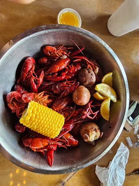 Overhead view of a Cajun crawfish boil with whole crawfish, corn on the cob, red potatoes, lemon wedges, and melted butter in a metal bowl.