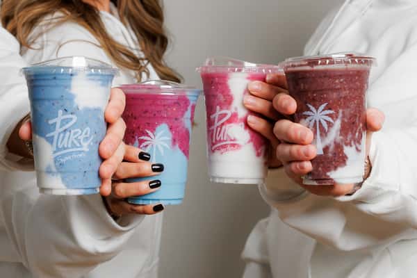 Close-up of hands holding three vibrant Pure Superfood smoothies in blue, magenta, and dark acai hues, swirled with creamy white yogurt.