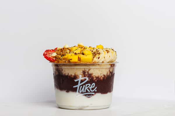 Layered acai bowl from Pure Superfood featuring creamy yogurt, rich acai blend, and oats, topped with fresh banana slices, mango chunks, strawberries, granola, flax seeds, and white chocolate chips.