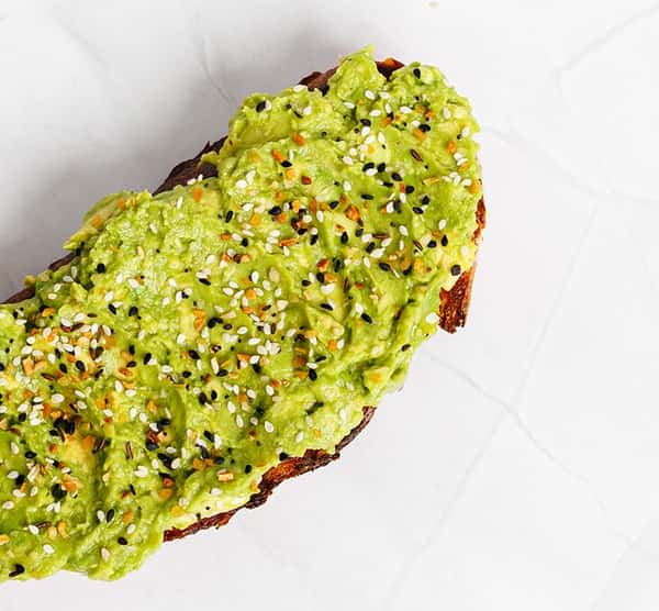 Crunchy multigrain toast topped with creamy smashed avocado and sprinkled with everything bagel seasoning, served on a white textured surface.