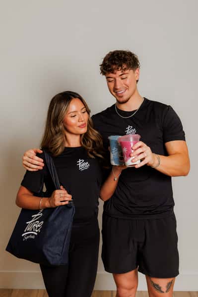 Smiling man and woman in matching Pure Superfoods outfits holding vibrant smoothies, showcasing branded healthy lifestyle with reusable tote.