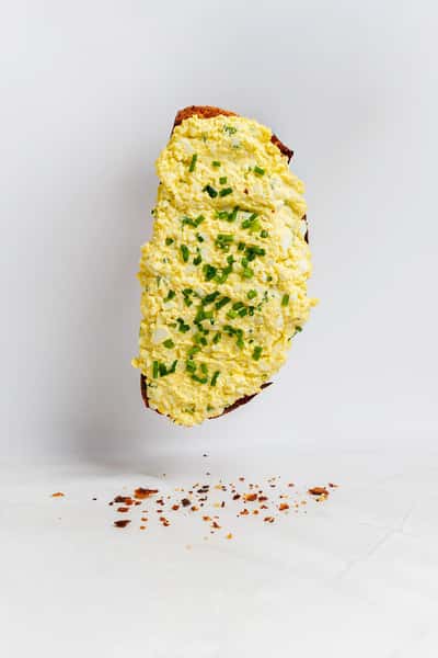 Rustic toast generously topped with creamy egg salad and garnished with chopped fresh chives, with scattered crumbs on a white background.