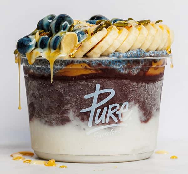 Close-up of a decadent Pure Superfood acai bowl layered with creamy yogurt, rich acai blend, and chia pudding, topped with sliced bananas, blueberries, almond slivers, pumpkin seeds, and a golden honey drizzle.