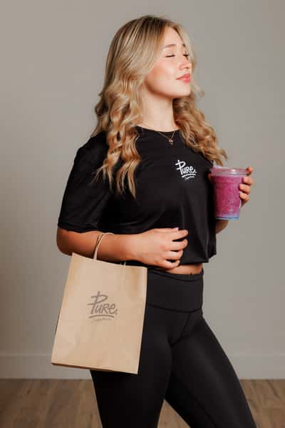 A woman in Pure Superfood athletic wear holds a bright purple smoothie and a branded takeaway bag, embodying a fresh, health-conscious lifestyle.