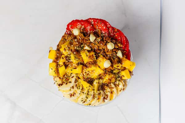 Top-down view of a vibrant Pure Superfood bowl topped with sliced strawberries, banana, fresh mango chunks, granola, macadamia nuts, flax seeds, hemp seeds, and a drizzle of nut butter.