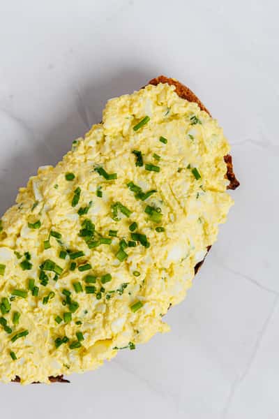 Open-faced egg salad toast topped with finely chopped chives on rustic whole grain bread, served on a minimalist white surface — a high-protein, café-style breakfast option.