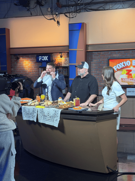 Behind-the-scenes Fox 10 Arizona live broadcast featuring Foley Ranch BBQ owners showcasing smoked barbecue, signature cocktails, and country bar favorites while the host samples food on air.