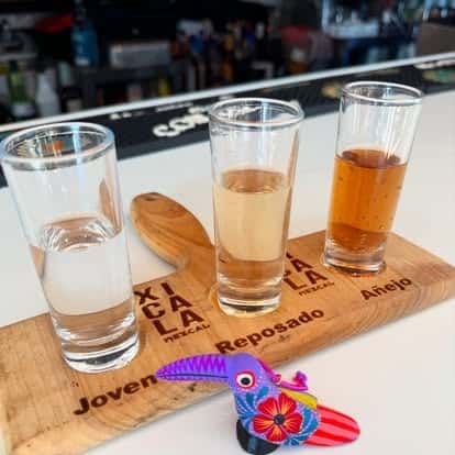 Mezcal flight featuring Joven, Reposado, and Añejo varieties served in shot glasses on a branded wooden tray with a colorful decorative toucan.