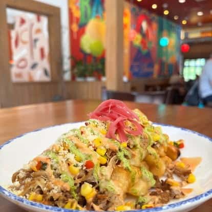 Loaded street fries topped with shredded pork, sweet corn, cotija cheese, pickled onions, and green drizzle sauce, served in a lively restaurant interior.