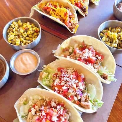 Platter of assorted soft tacos with seasoned meat, fresh toppings, and crumbled cheese, paired with roasted corn, chipotle mayo, and salsa bowls.