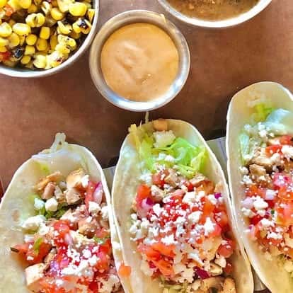 Trio of grilled chicken tacos with shredded lettuce, diced tomatoes, cotija cheese, and soft flour tortillas, served with creamy dipping sauce and roasted corn.