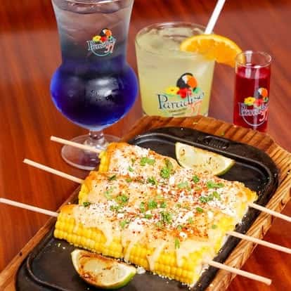 Grilled Mexican street corn skewers with cotija cheese, chili, and cilantro, served with vibrant tropical cocktails including a blue margarita and citrus punch.
