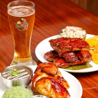 Barbecue ribs and roasted chicken served with cilantro rice, creamy coleslaw, and crispy plantain chips, paired with a chilled draft beer — bold Latin American barbecue feast.