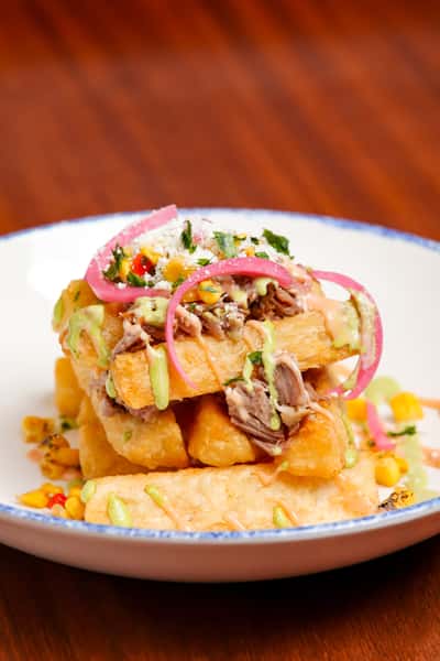 Loaded yucca fries stacked and topped with tender shredded beef, pickled red onions, roasted corn, cotija cheese, and drizzles of creamy avocado and chipotle sauces.