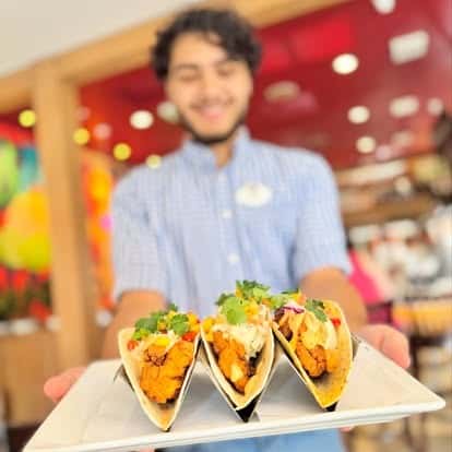 Smiling server presenting a trio of crispy chicken tacos topped with fresh cilantro, avocado crema, and pickled onions in a vibrant restaurant setting.
