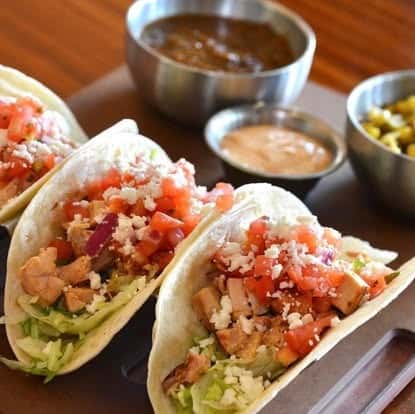 Freshly prepared tacos filled with diced chicken, lettuce, tomatoes, and crumbled cheese, served with roasted corn, spicy salsa, and creamy chipotle sauce.