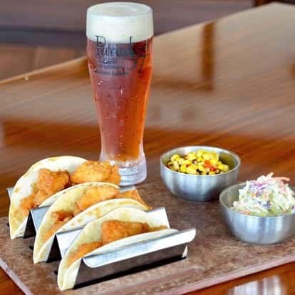 Crispy fried fish tacos served in soft tortillas with a side of roasted corn and creamy coleslaw, paired with a cold glass of amber beer.