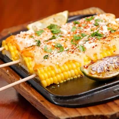 Elote-style grilled corn on the cob with melted cheese, chili powder, cotija, and chopped cilantro, served with grilled lime wedges on a sizzling plate.