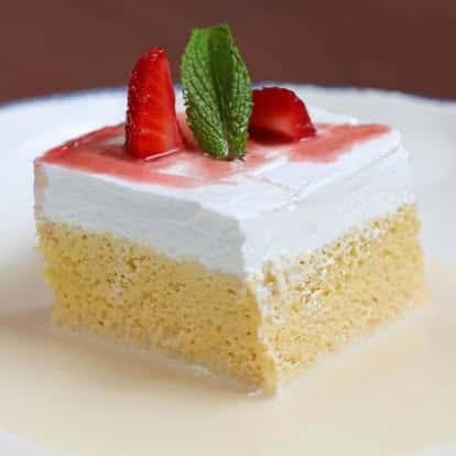 Moist tres leches cake topped with whipped cream, fresh strawberries, and mint leaves, sitting in a pool of sweet milk sauce for a classic Latin dessert.