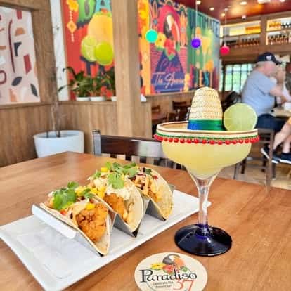 Trio of crispy chicken tacos topped with fresh cilantro and creamy sauce, served alongside a festive margarita garnished with a lime and mini sombrero in a colorful restaurant setting.