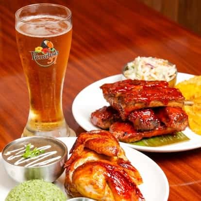 Feast platter featuring roasted chicken, BBQ-glazed skewers, green rice, black beans, plantain chips, and coleslaw, served with a chilled draft beer.