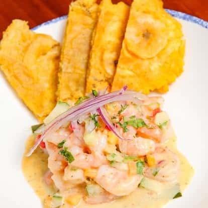 Creamy shrimp ceviche with diced cucumber, red onions, and cilantro, served with crispy golden tostones on a white and blue-rimmed plate.