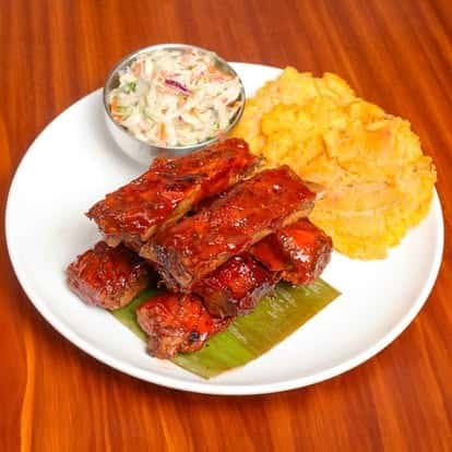 Juicy barbecue pork ribs served with creamy coleslaw and mashed sweet plantains on a white plate over a banana leaf base.
