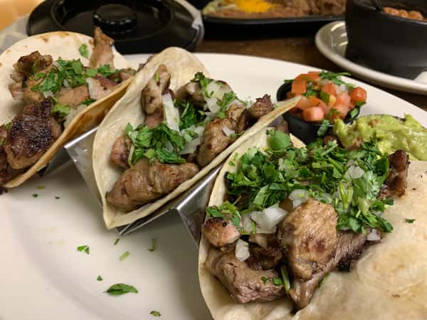 Three carne asada tacos topped with chopped onions and cilantro, served with guacamole and pico de gallo on the side.