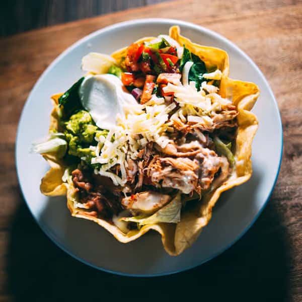Taco salad served in a crispy tortilla bowl with shredded chicken, cheese, guacamole, sour cream, beans, and pico de gallo.
