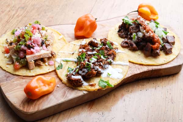 Three street tacos with grilled chicken, chorizo, and carne asada on corn tortillas, garnished with salsa, onions, and cilantro.