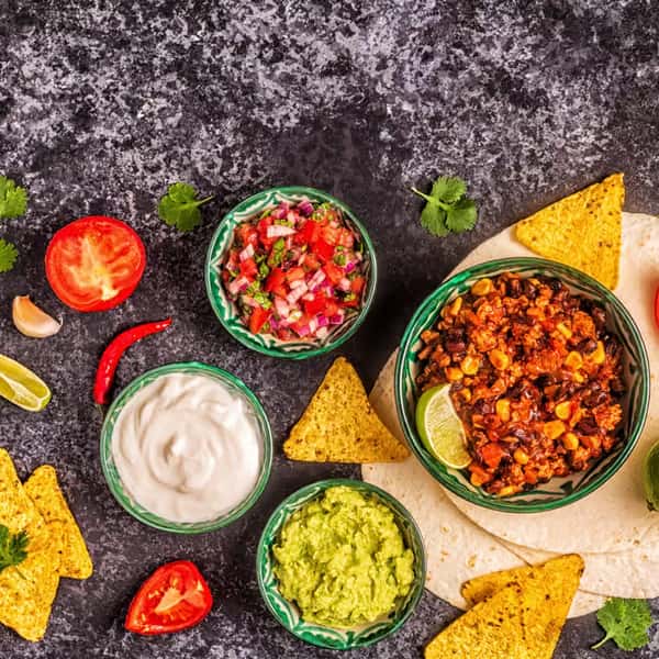 Mexican appetizer spread with tortilla chips, guacamole, sour cream, pico de gallo, and chili with corn in festive bowls.