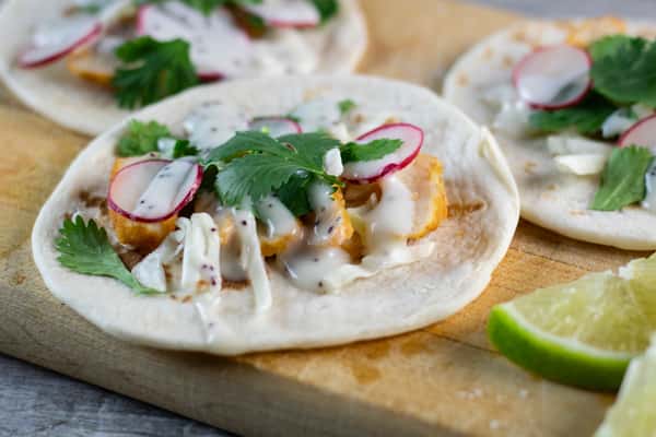 Fish tacos topped with sliced radish, cabbage, cilantro, and creamy dressing on soft tortillas with lime wedges on the side.