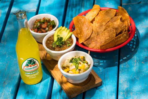 Basket of tortilla chips with pico de gallo, salsa, pineapple salsa, and Jarritos pineapple soda on a blue wooden table.