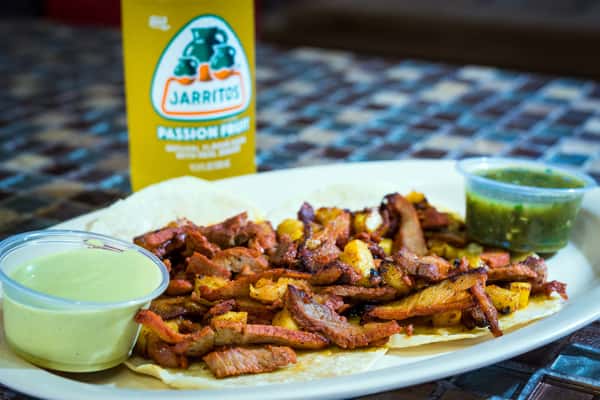 Tacos al pastor served open-faced on corn tortillas, topped with grilled marinated pork and chunks of pineapple. Accompanied by creamy avocado salsa, green salsa, and a bottle of Jarritos passion fruit soda.