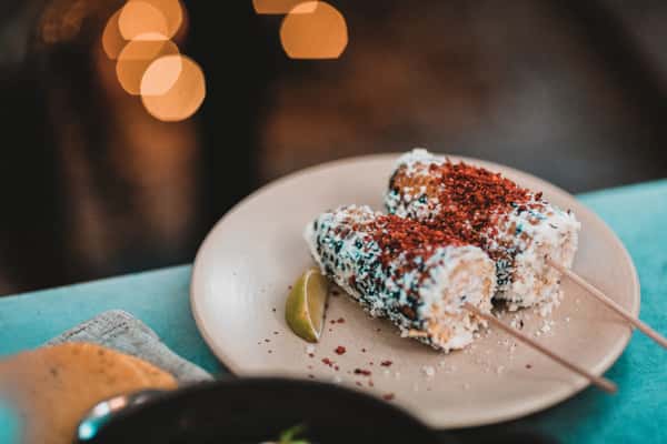 Mexican street corn on sticks topped with cotija cheese and chili powder, served with a lime wedge on a beige plate.