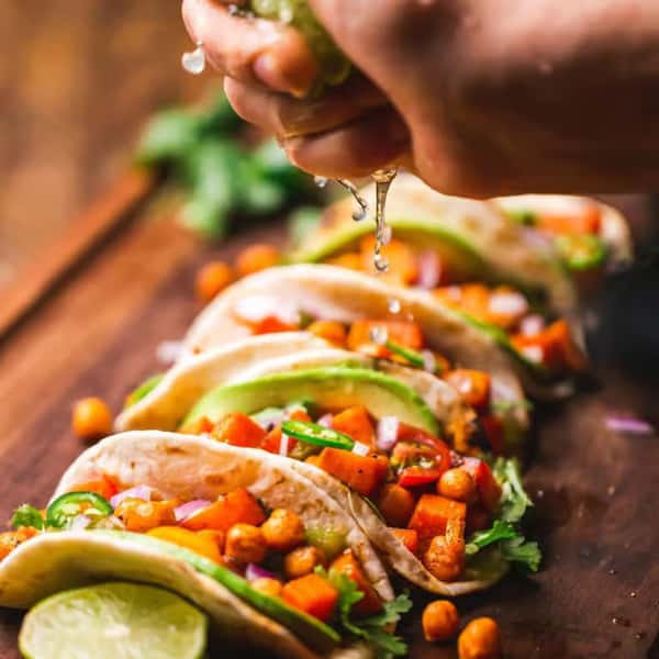 Soft tacos filled with chickpeas, diced vegetables, and avocado slices being topped with freshly squeezed lime juice.