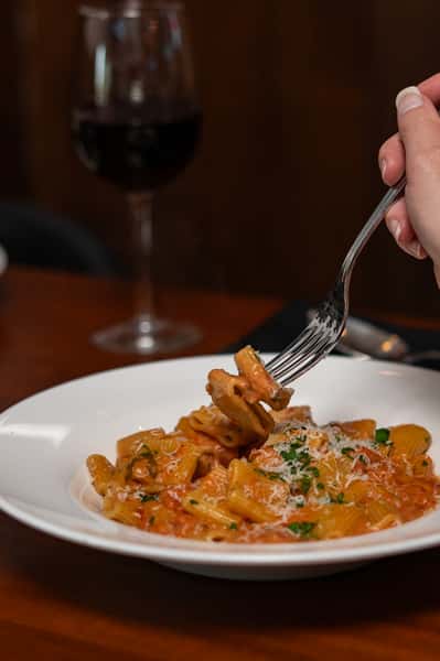 Fork lifting creamy rigatoni pasta in tomato sauce, topped with grated cheese and parsley, with a glass of red wine in background.