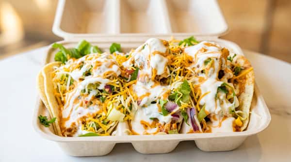 Tacos in a biodegradable container, topped with lettuce, shredded cheese, white sour cream, and orange sauce.