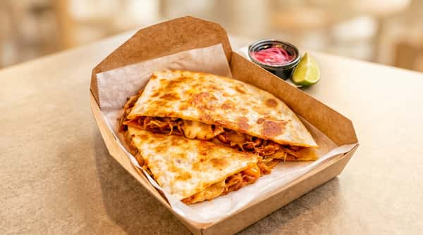 Two chicken and cheese quesadillas in a cardboard container with a side of pickled red onions and a lime wedge.