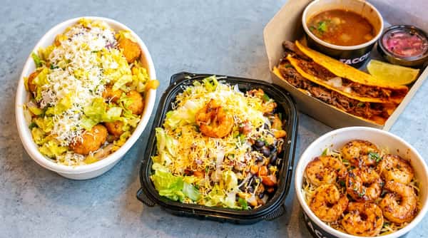 Two salad bowls with cheese and lettuce, next to a taco box with birria tacos, consommé, and lime.