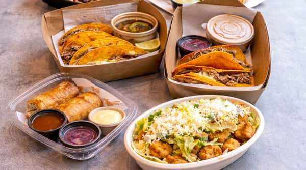 Birria tacos with consommé and lime, a salad with fried chicken and cheese, and egg rolls with dipping sauces.
