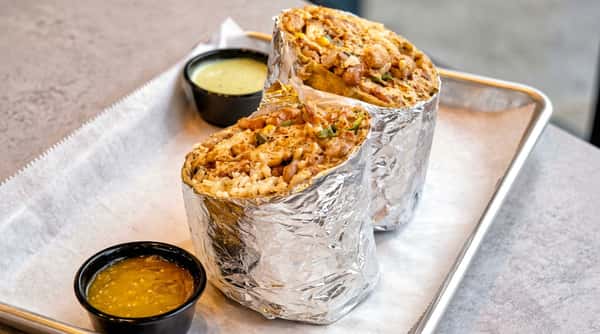 Two foil-wrapped burritos with visible rice, meat, and corn filling, served with green and yellow sauces.