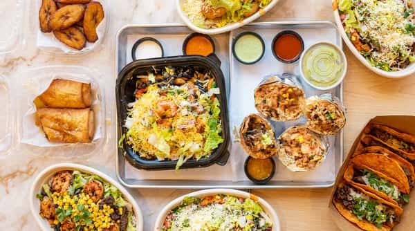 Top-down view of various Mexican dishes: shrimp salad, several burritos, tacos, spring rolls, and dipping sauces.