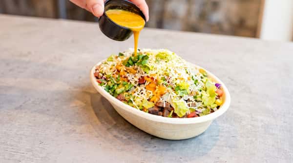 Burrito bowl with beef, cheese, lettuce, and salsa, with sauce being poured over it.