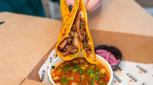 Hand holding two birria tacos with shredded meat, dipped in a rich red consommé, alongside pickled red onions.