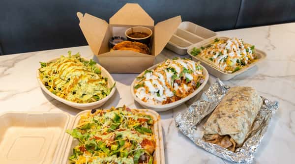 Assortment of take-out Mexican food including birria tacos, burrito bowls with various toppings, and a foil-wrapped burrito.